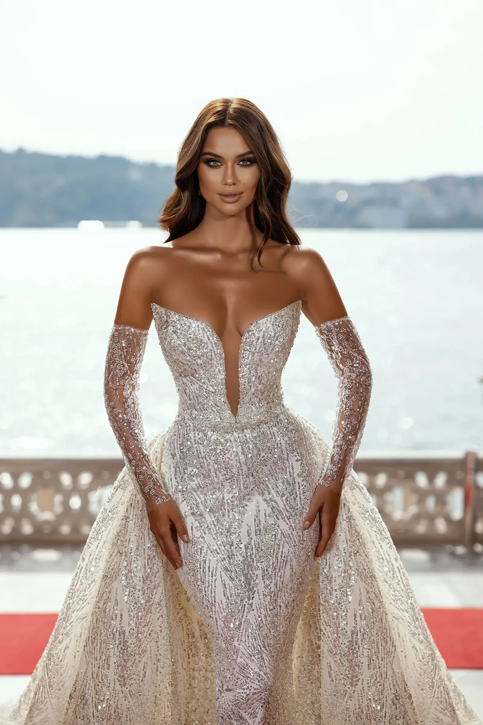 A woman in a sparkling off-shoulder gown with intricate beading stands confidently near a sunlit waterfront. Her expression is poised and elegant.