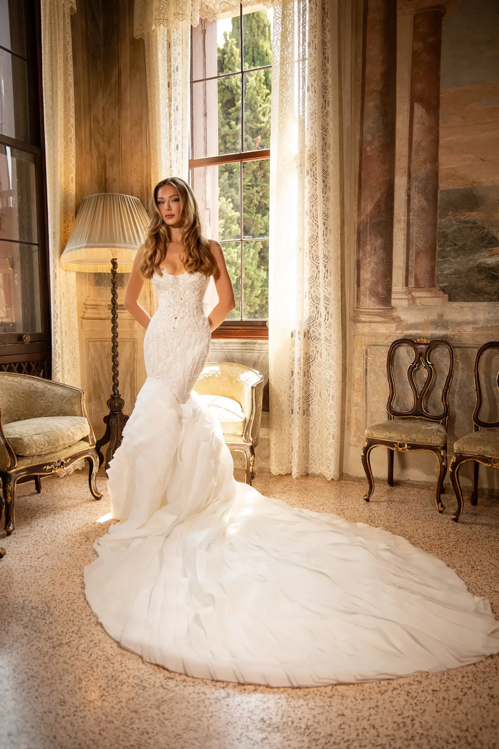 A woman in a white wedding dress stands gracefully in a sunlit room with vintage decor, including a lamp and ornate chairs.