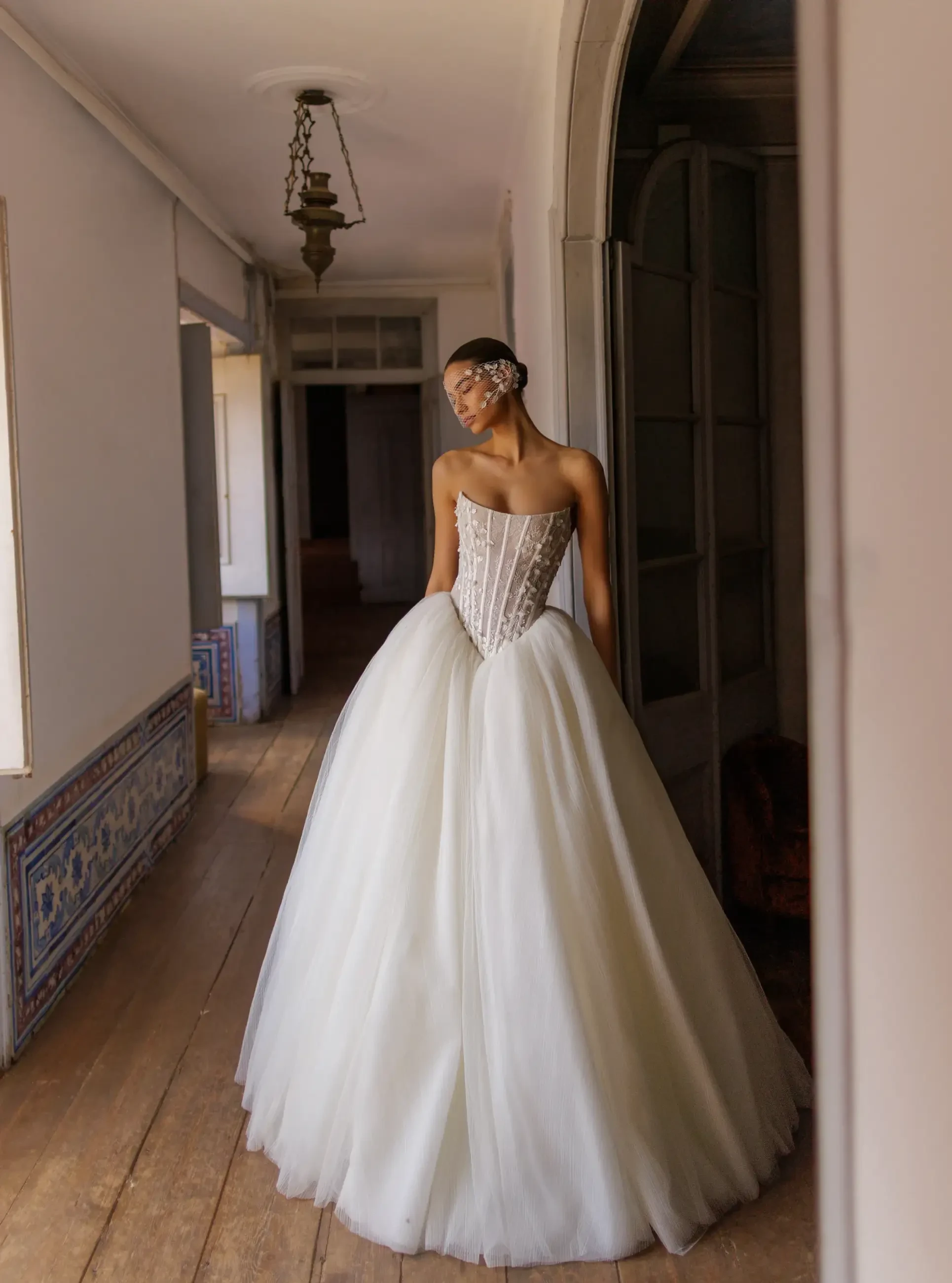 A woman in a beautiful white gown with a corset top and a full, flowing skirt stands gracefully in an elegant hallway.