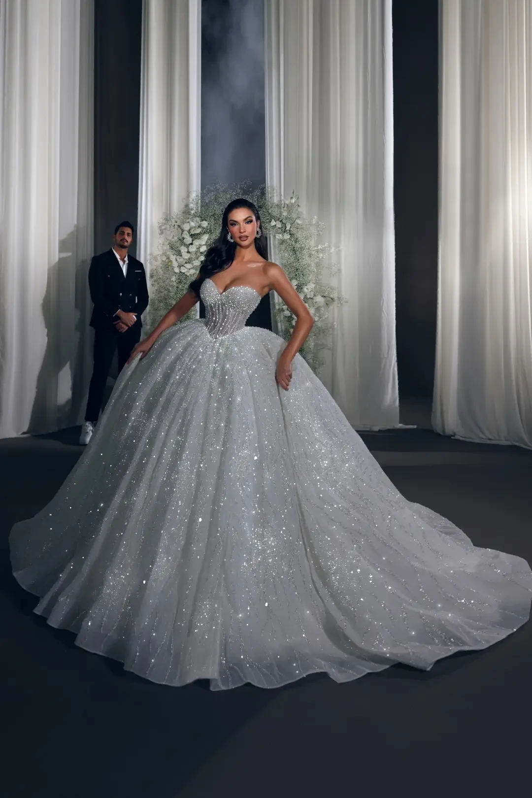 A woman in a sparkling, voluminous white wedding dress poses confidently in front of white draped curtains. A man in a suit stands behind her, adding elegance to the scene.