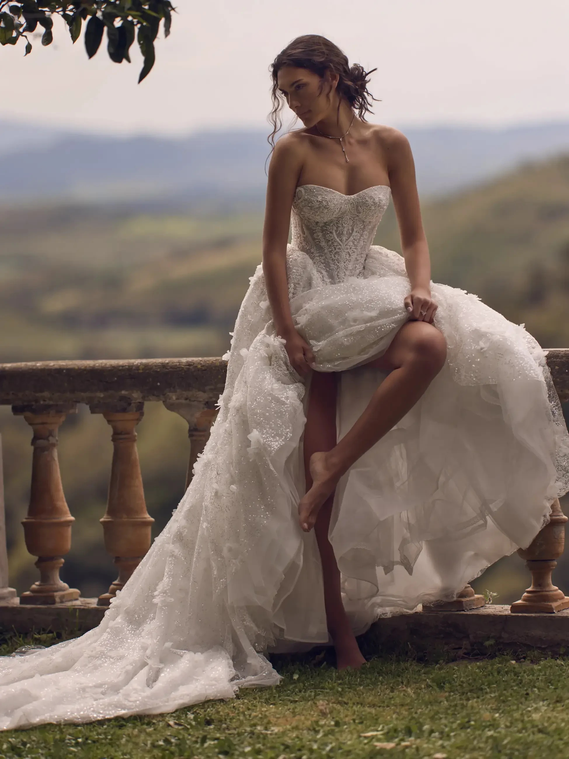 A woman in a strapless wedding dress poses gracefully, holding up the skirt with one hand while resting her other foot on a stone railing. The background features scenic hills and greenery.