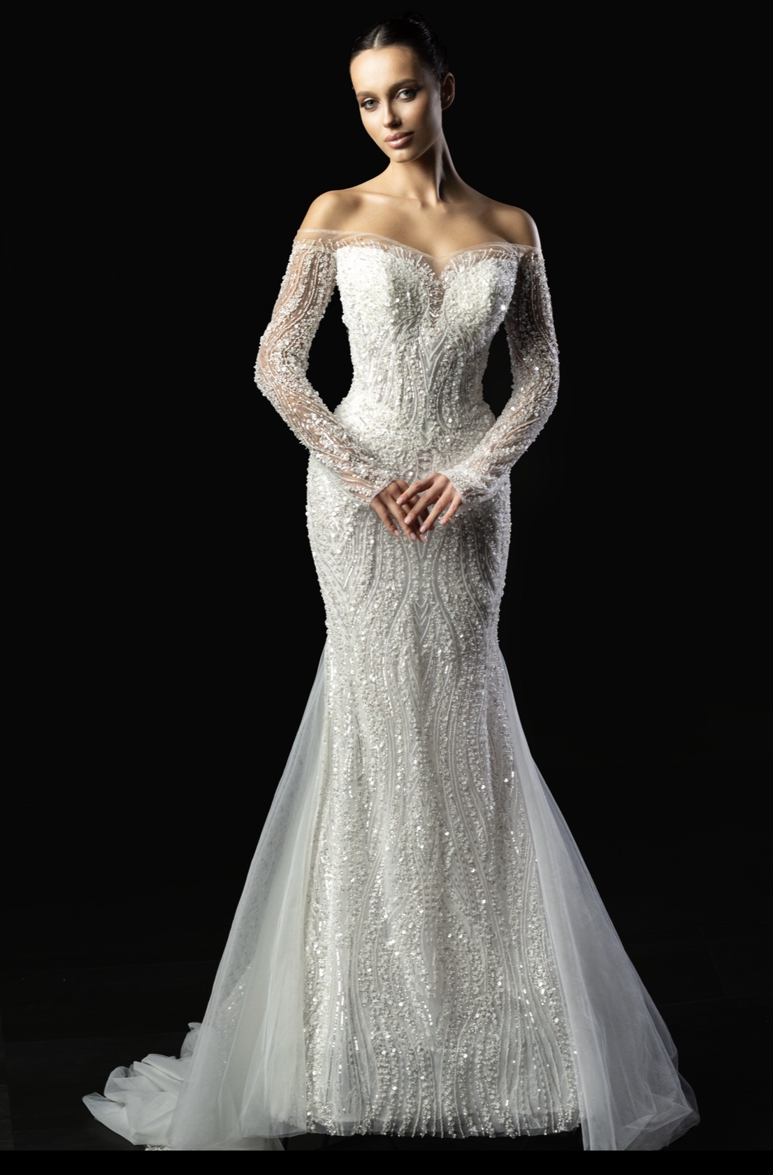 A model wearing an elegant off-shoulder wedding gown with intricate beadwork and long sheer sleeves, set against a dark background.