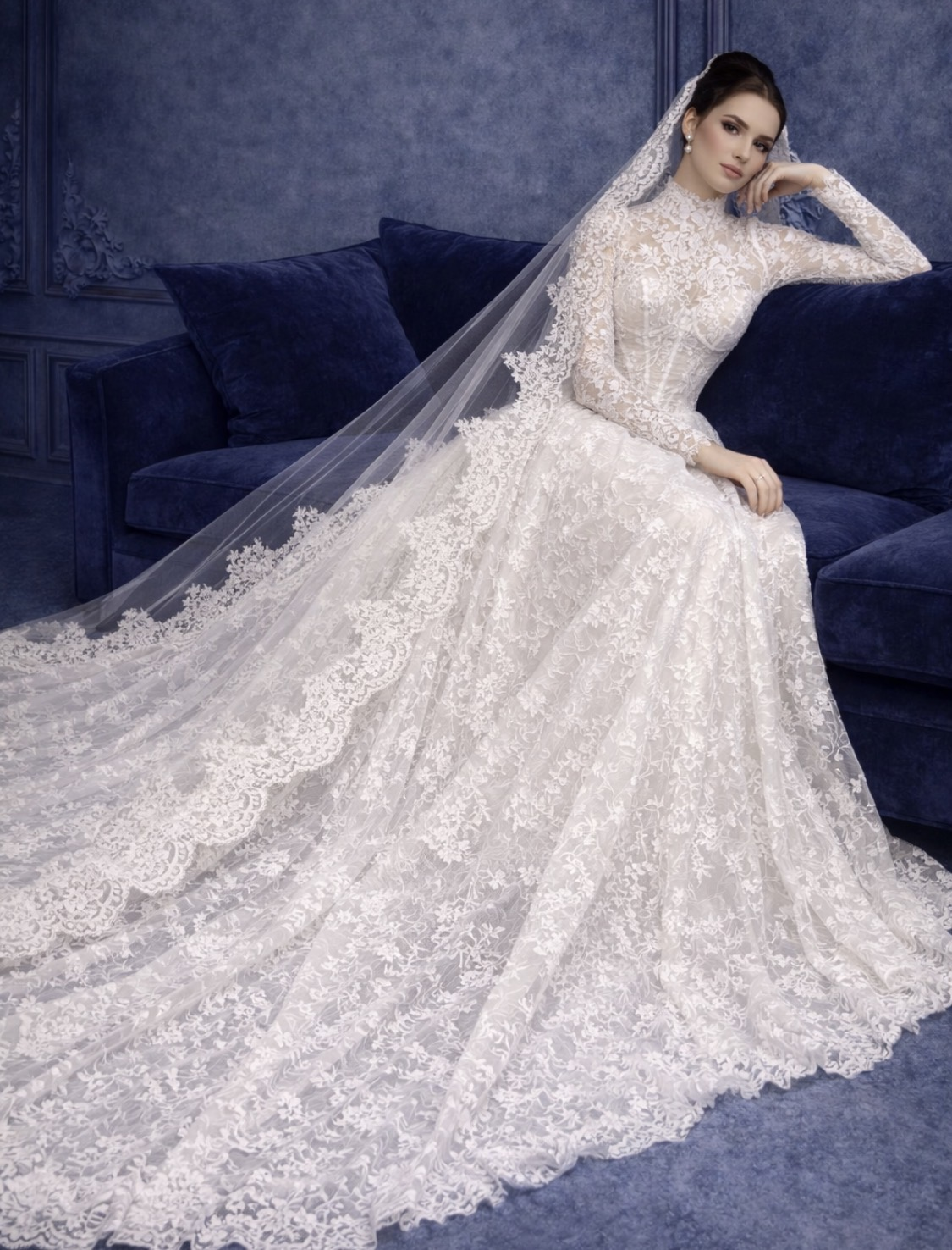A woman wearing a long, elegant white lace wedding dress with intricate detailing, sitting on a blue velvet couch. She has long dark hair and is wearing a matching lace veil. The background features blue decor.