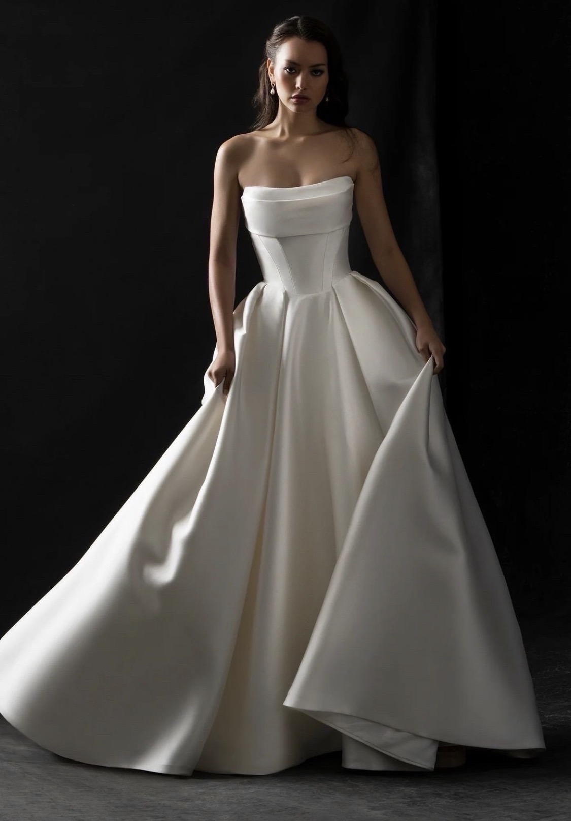 A model wearing a strapless, flowing white gown, posing against a dark backdrop.