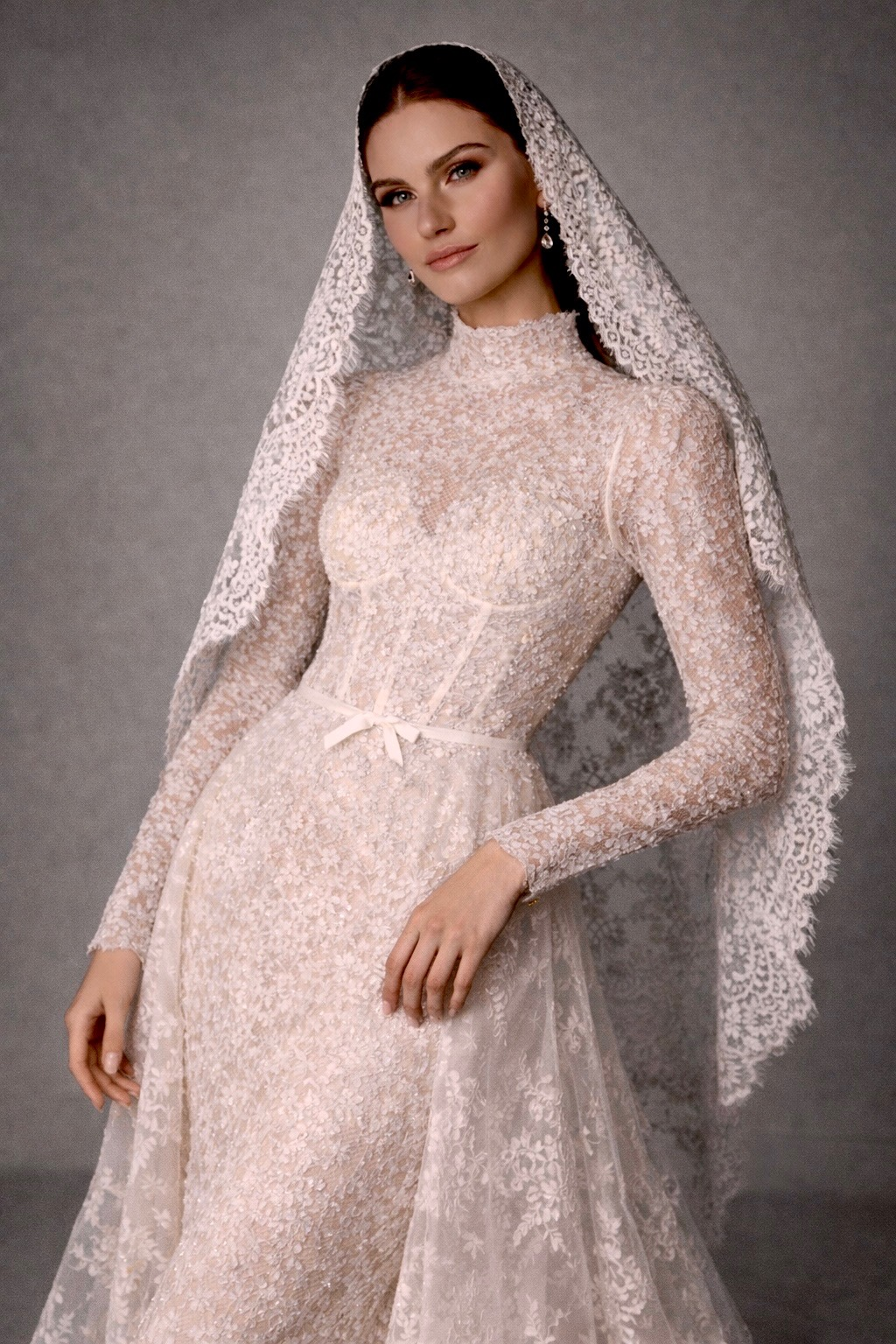 A model wearing a delicate lace wedding dress with long sleeves and a matching lace veil, posing against a neutral background.