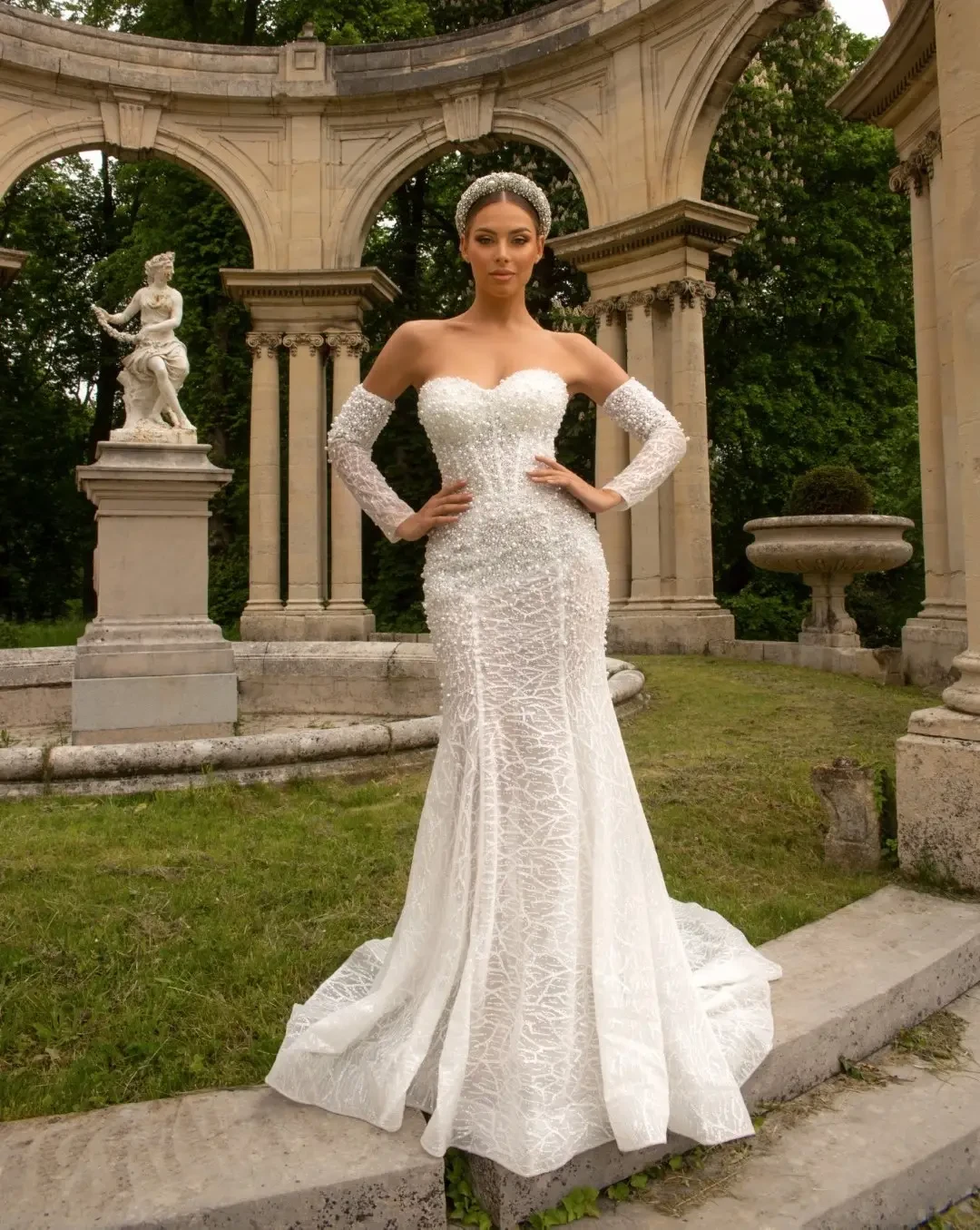 Woman in an elegant lace wedding dress stands confidently in front of a neoclassical stone arch, surrounded by greenery. The scene is serene and majestic.