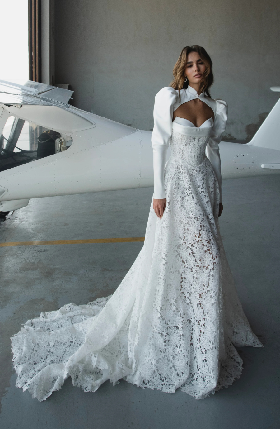 Model is wearing a fit-and-flare lace bridal gown with a high-neck bolero, long sleeves, and a corset bodice.