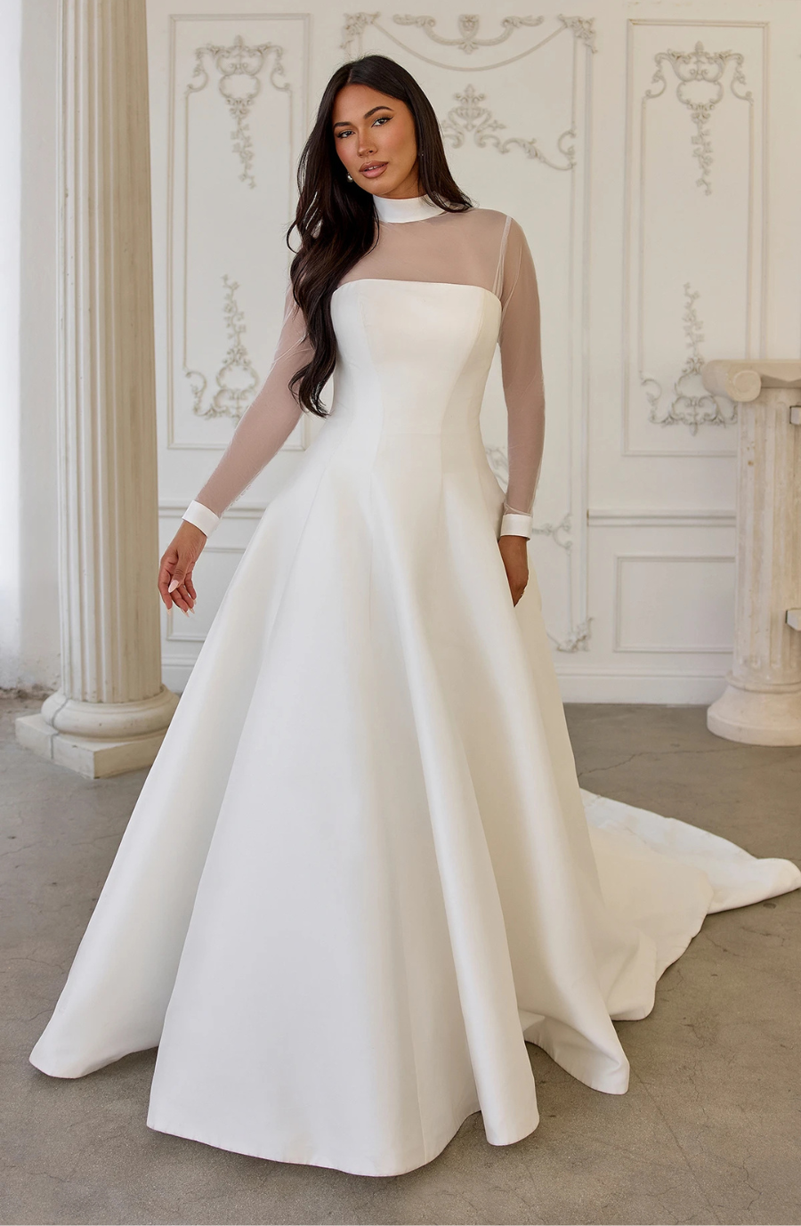 Model is wearing a modest ball gown with sheer illusion long sleeves, a high neckline, smooth fabric, and delicate cuffs at the neck and wrists.