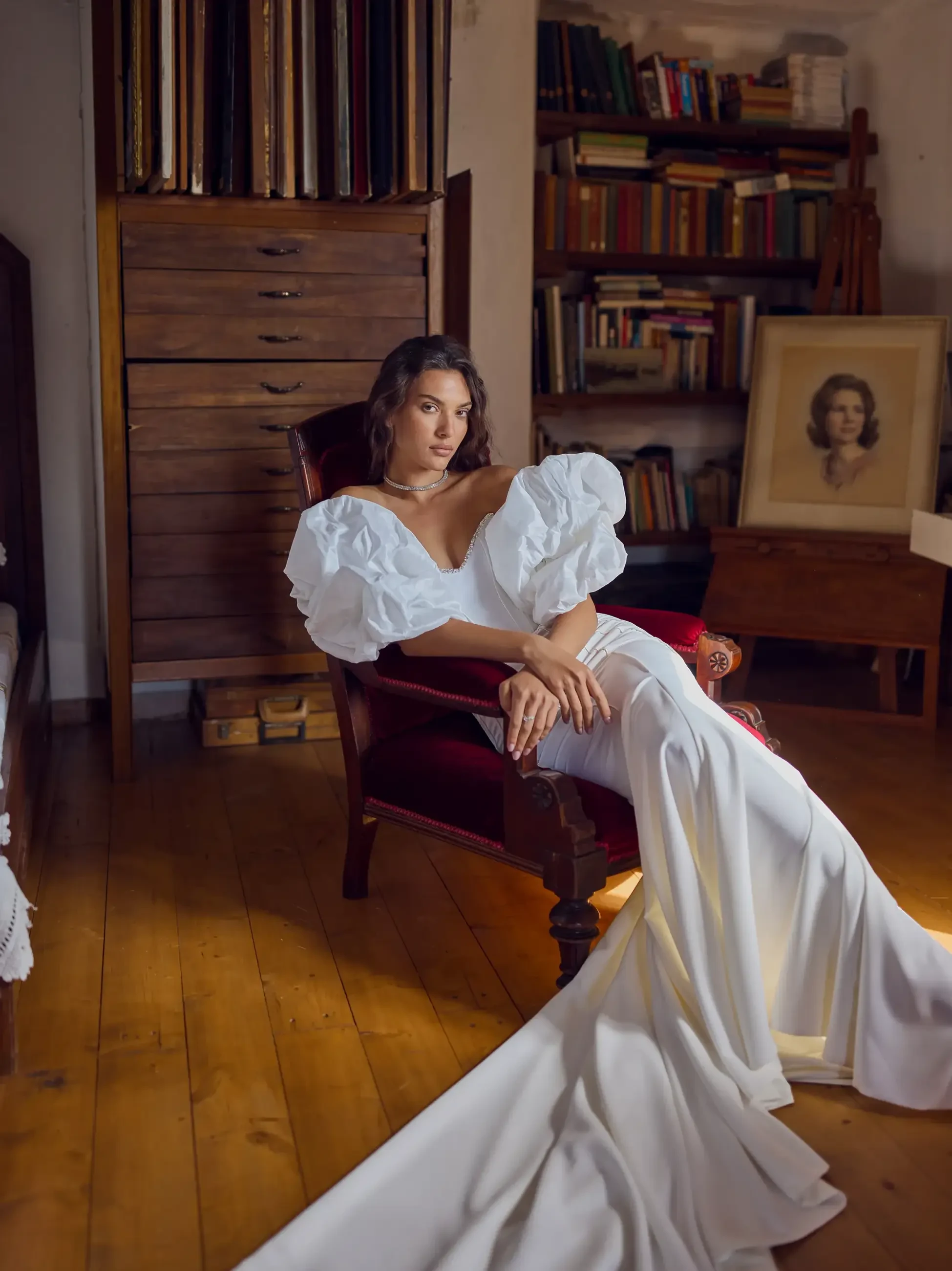A woman in a stylish white dress with puffed sleeves poses elegantly on a chair in a room filled with books.